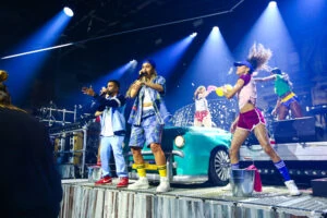 Vocalists and dancers performing around a turquoise prop car under blue stage lighting.